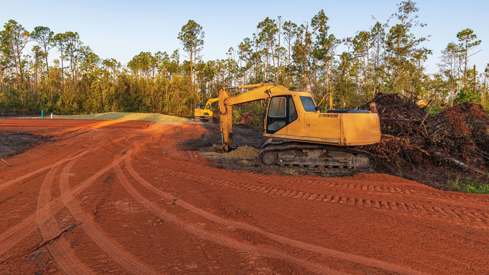 Yellow excavator on red dirt clearing land near pine forest