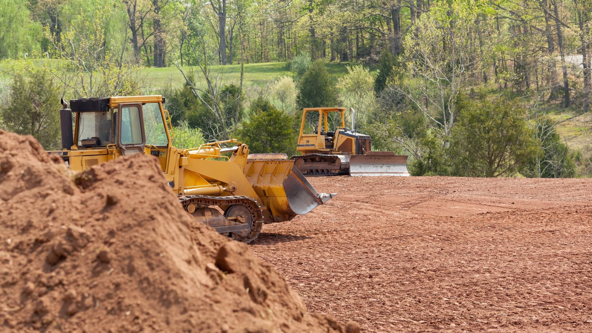 Yellow bulldozers clearing land in wooded area with dirt piles